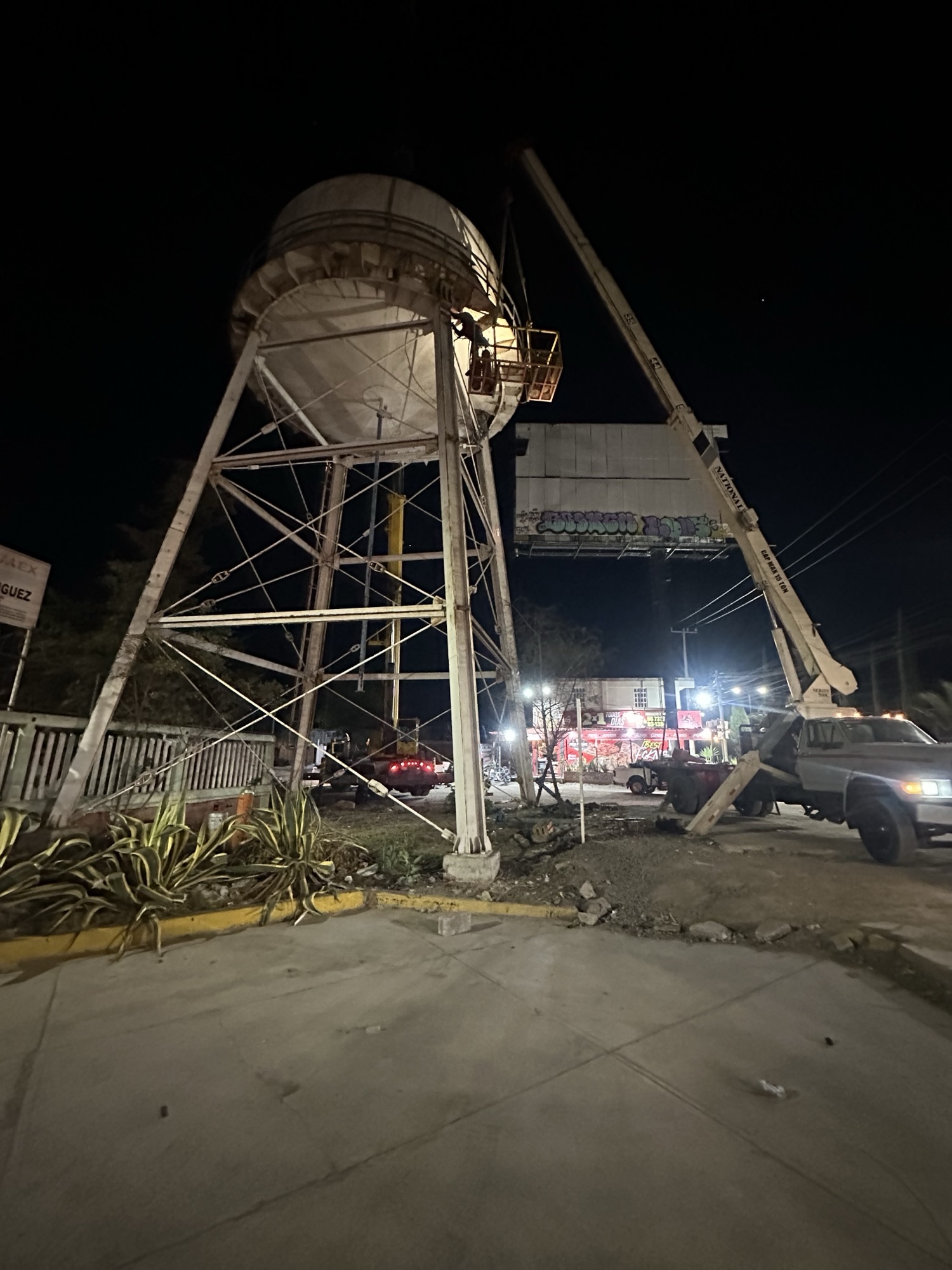 Desmontaje nocturno de un tanque de acero sobre torre estructural con grúa telescópica y canastilla de personal